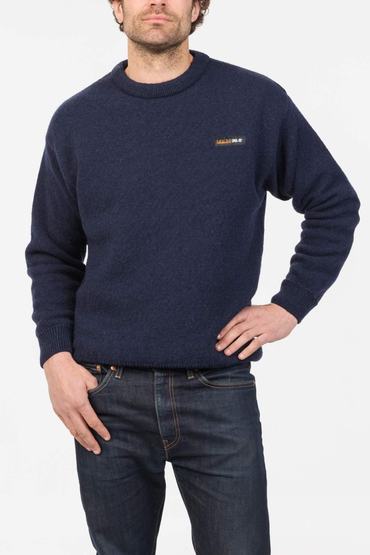 A man wearing a dark blue MKM Tasman Dual Layer Possum & Merino Crew sweater with a small chest logo and blue jeans stands against a white background, one hand in his pocket and the other resting on his hip.