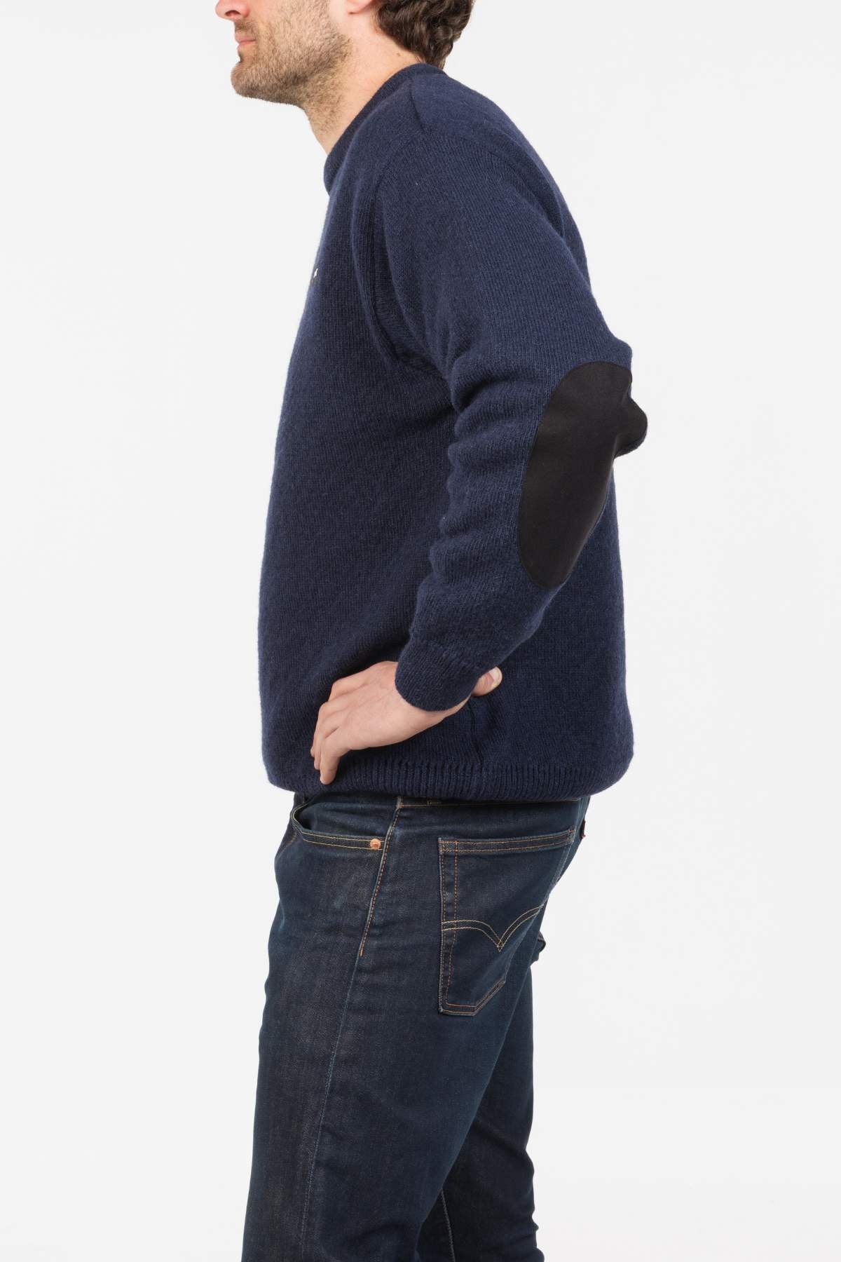 A man wearing an MKM Tasman Dual Layer Possum & Merino Crew sweater in dark blue with black elbow patches and blue jeans stands in profile, hand on hip, against a plain white background.