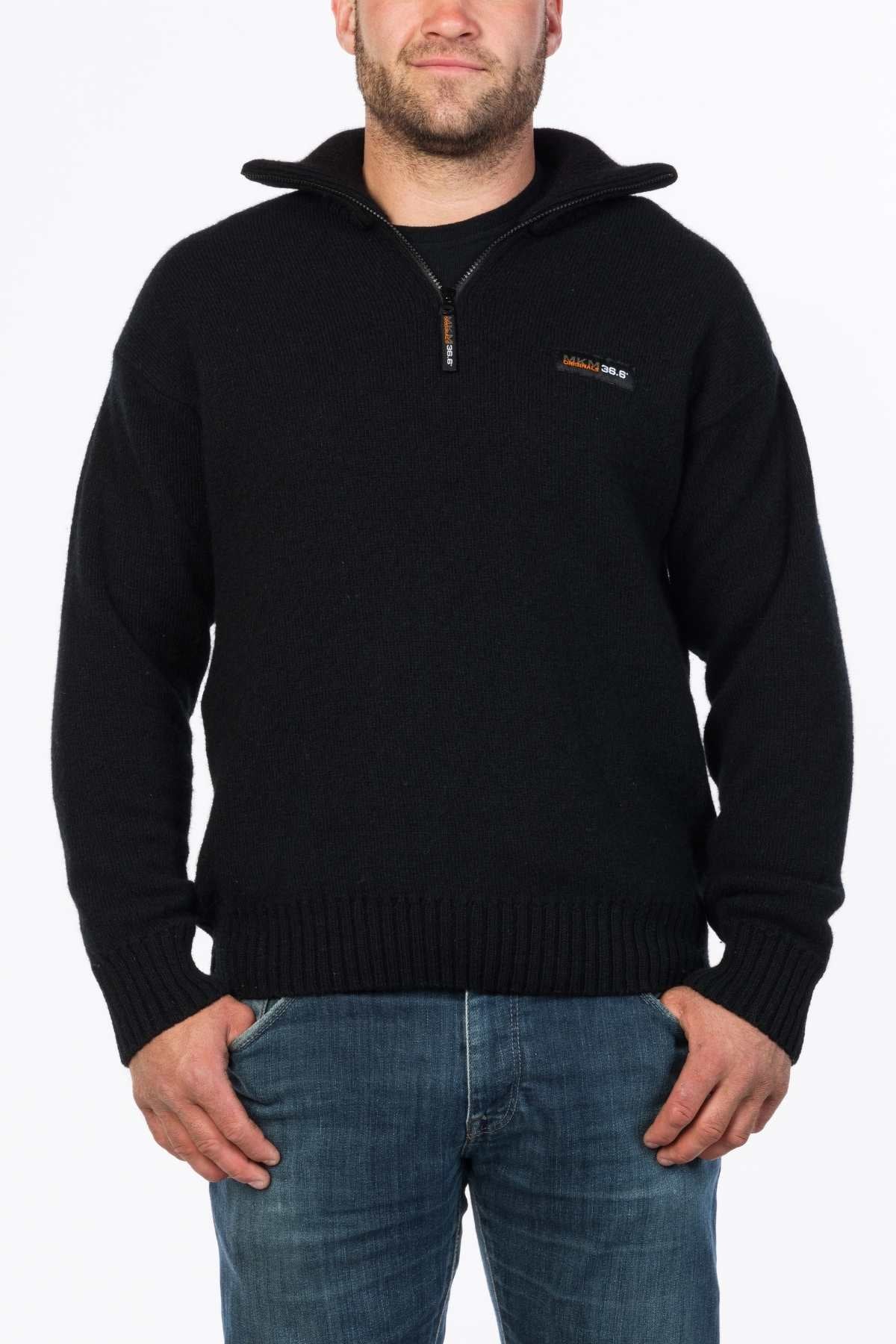 Dressed in the MKM Tasman Dual Layer Possum & Merino Knit, a person stands against a white background. The cozy black sweater, adorned with a small chest logo, is paired with blue jeans as they clasp their hands at the front.