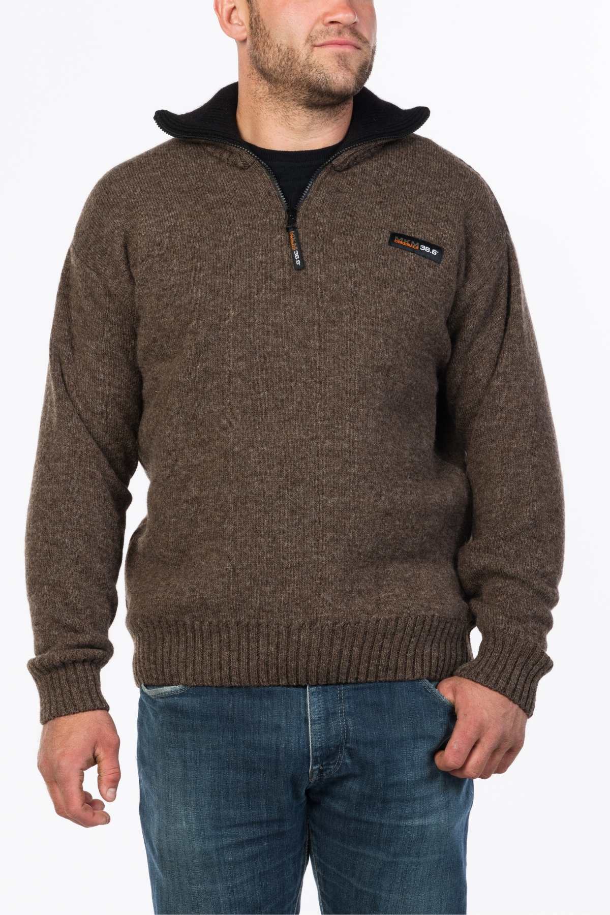 A man in an MKM Tasman Dual Layer Possum & Merino Knit, featuring a brown zip-up design with a chest logo, stands against a plain white background wearing blue jeans.