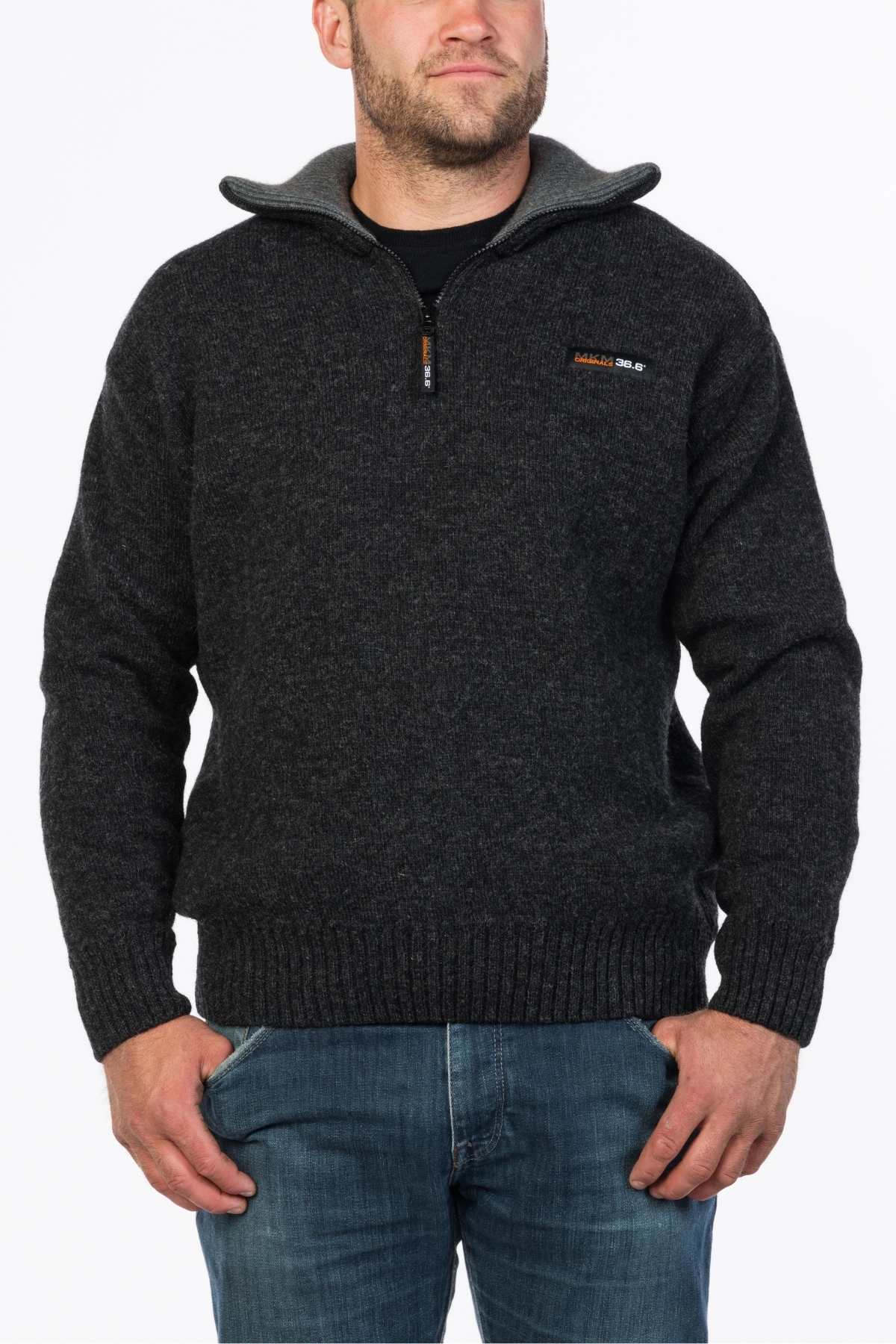 A man wearing an MKM Tasman Dual Layer Possum & Merino Knit, a dark gray zip-up sweater made of merino superfine wool with a high collar, stands with his left hand in the pocket of his blue jeans. The sweater features a small orange and white logo on the left chest. The background is plain white.