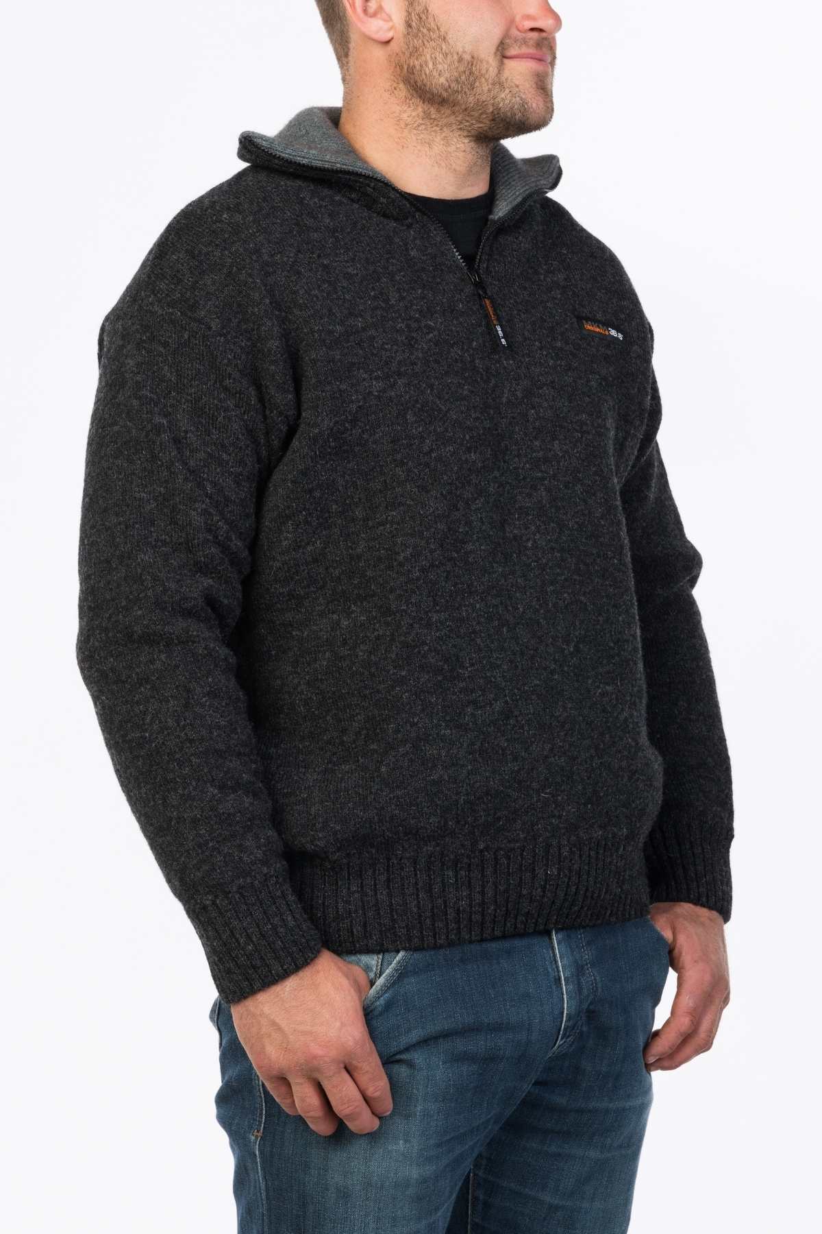A man is wearing the dark gray MKM Tasman Dual Layer Possum & Merino Knit sweater, which features a quarter-zip collar and long sleeves, paired with blue jeans. Made from superfine merino wool blended with possum fiber, he stands against a plain white background with one hand in his pocket and the other arm relaxed at his side, facing slightly to the right.