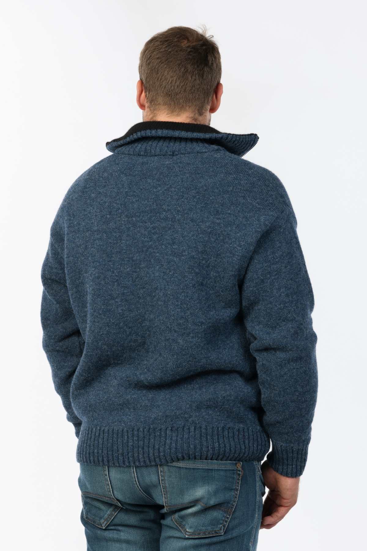 A man facing away from the camera is wearing a high-collared MKM Tasman Dual Layer Possum & Merino Knit blue sweater, paired with blue jeans, against a plain white background.