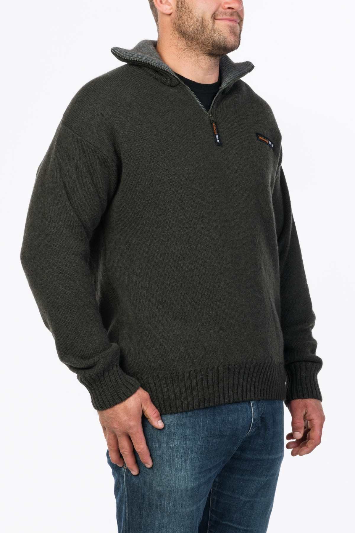 A person wearing an MKM Tasman Dual Layer Possum & Merino Knit in dark green, featuring a quarter-zip with ribbed cuffs and hem, matched with blue jeans. The sweater shows a distinct zipper and a small logo on the left chest. Made in New Zealand, the outfit contrasts against the plain white background.