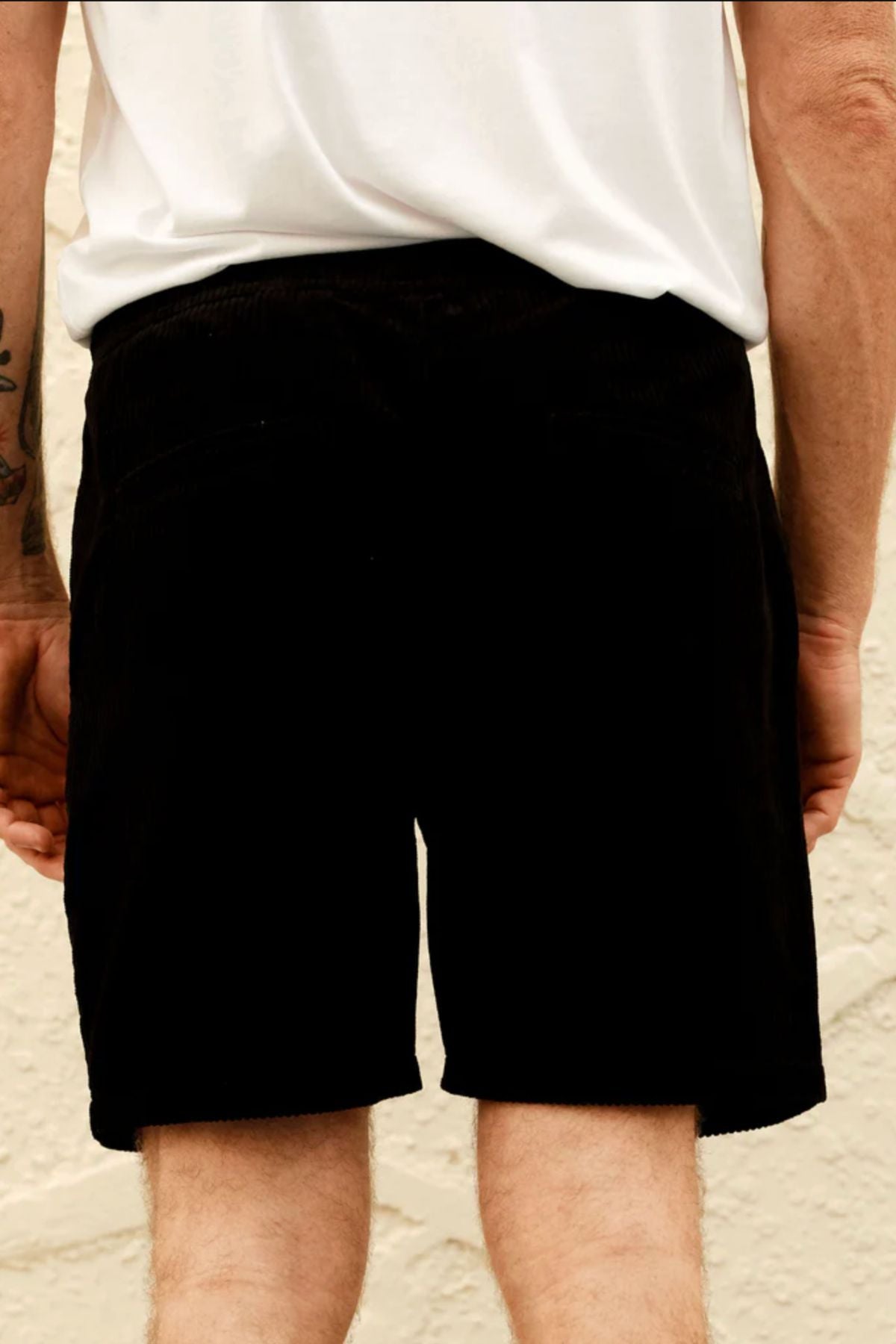 Someone wearing a white t-shirt and Mr Simple Burbank Cord Short in black stands with their back to the camera, arms at their sides, against a light textured wall.