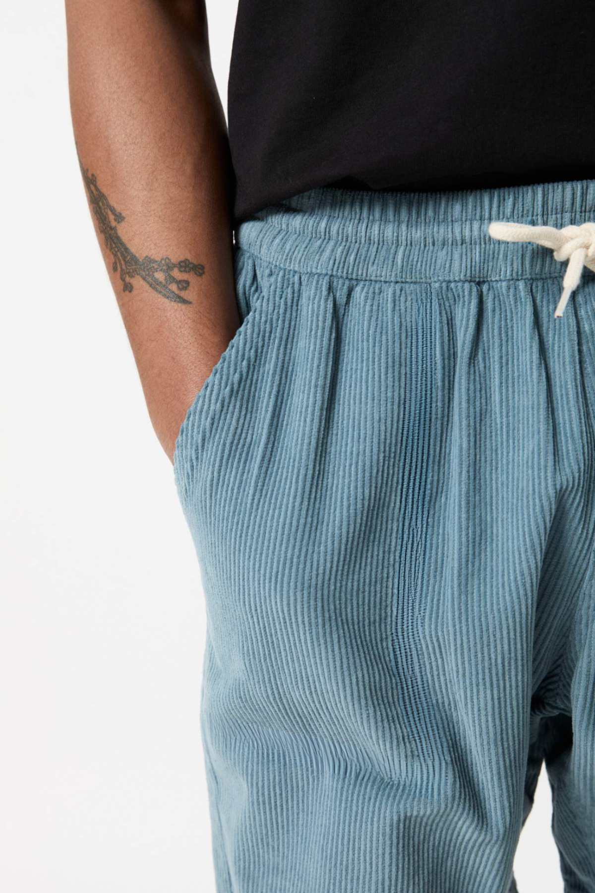 A close-up of a person wearing the light blue Mr Simple Burbank Cord Shorts, featuring an elastic waistband and a white drawstring. The person has a tattoo of a branch on their forearm and is wearing a black shirt, with their hand partially in the shorts pocket—a perfect look for a casual outing.
