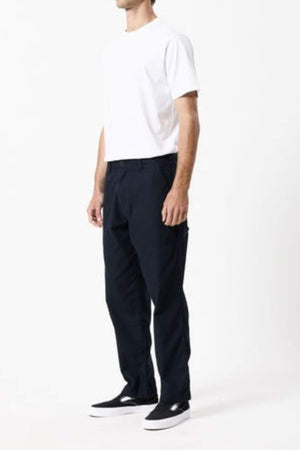 A person stands against a plain background wearing a white short-sleeve t-shirt, Mr Simple Carpenter Pant with a relaxed workwear-inspired design, and black slip-on shoes with white soles. The person's face is not visible.