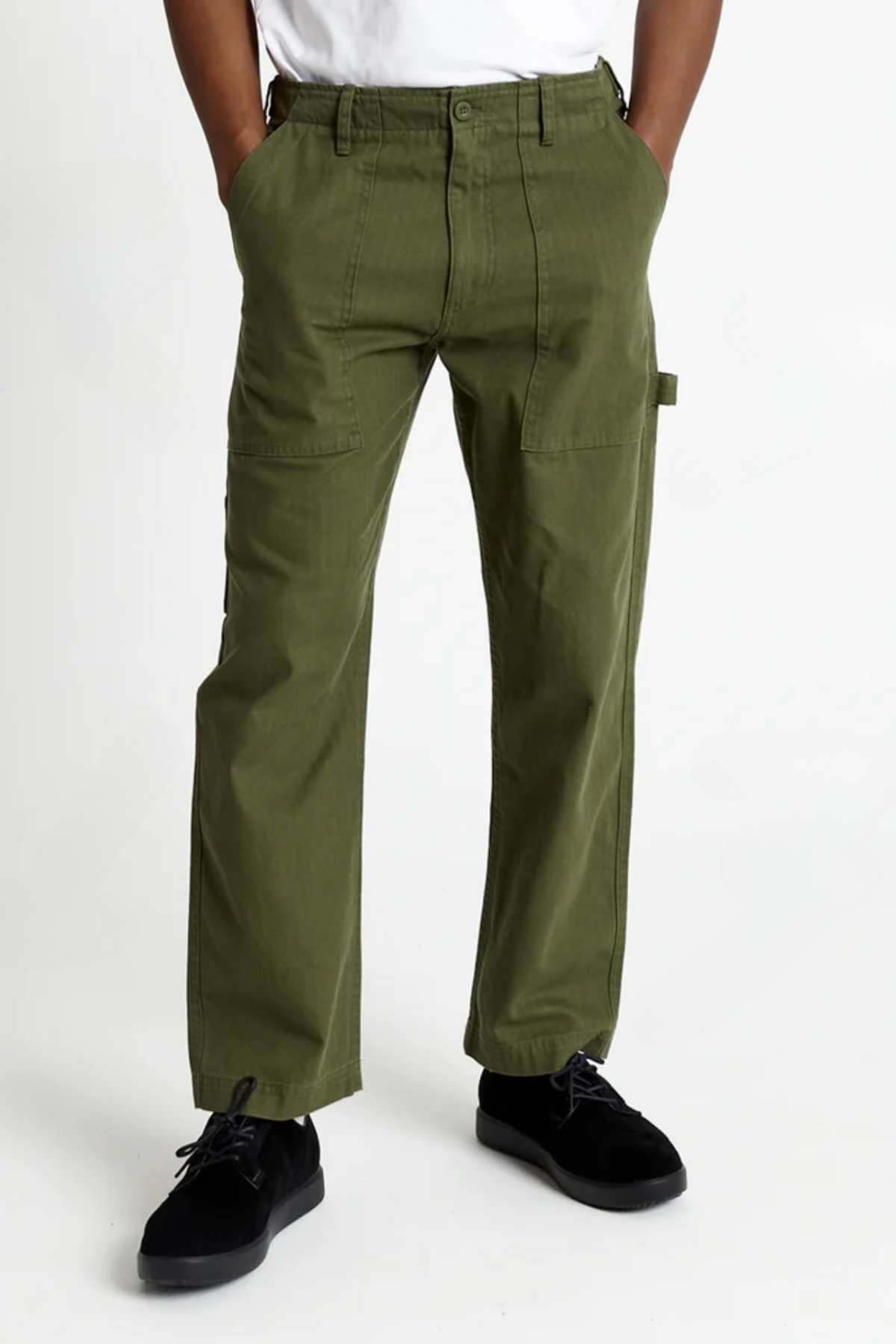 A person stands with hands in pockets, wearing the Mr Simple Carpenter Pant in olive green and black shoes, against a plain white background. Only the lower half of their body is visible.