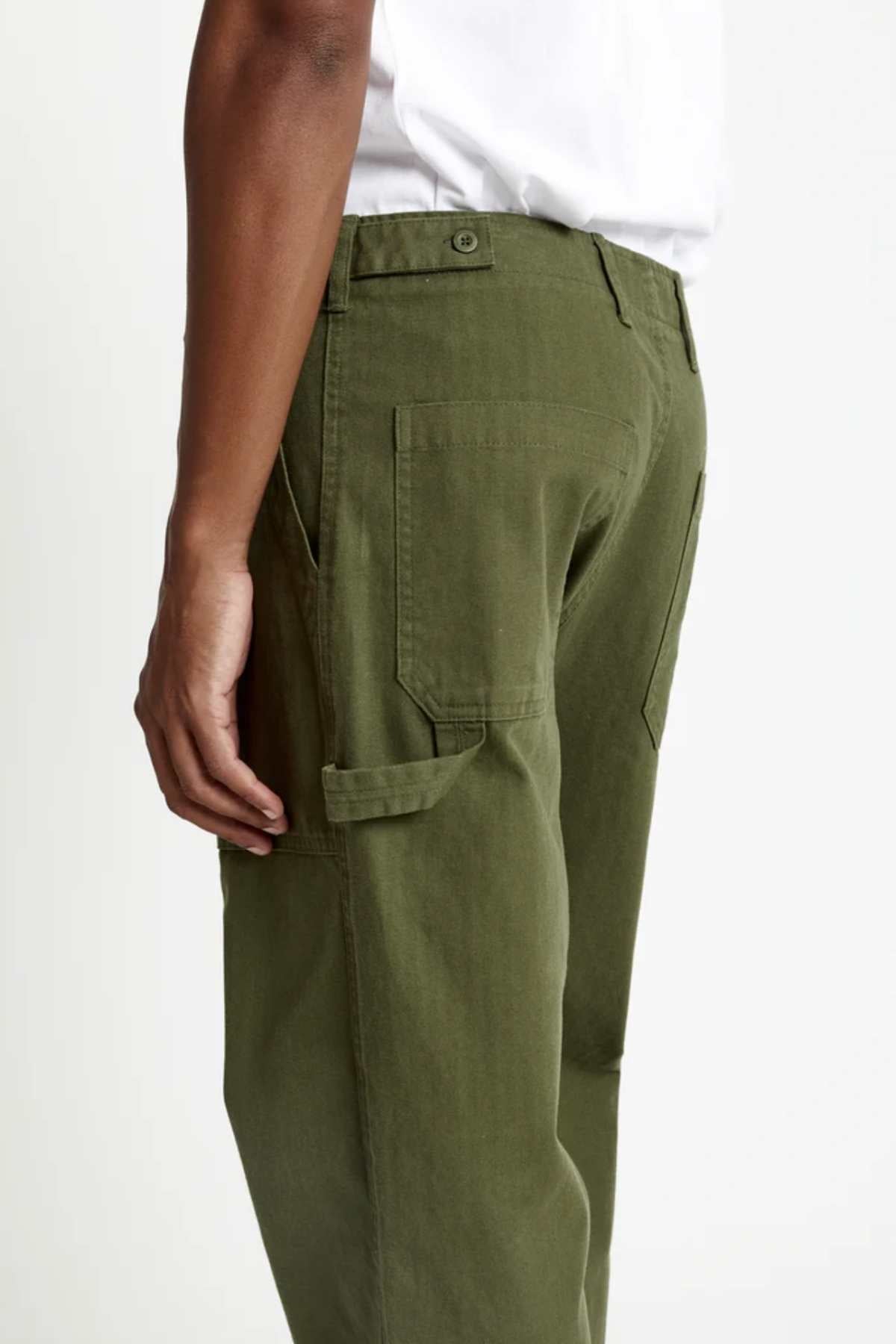 A person is shown from the side, waist down, wearing a white shirt and Mr Simple Carpenter Pant in olive green with a workwear-inspired design, one hand relaxed by their side.