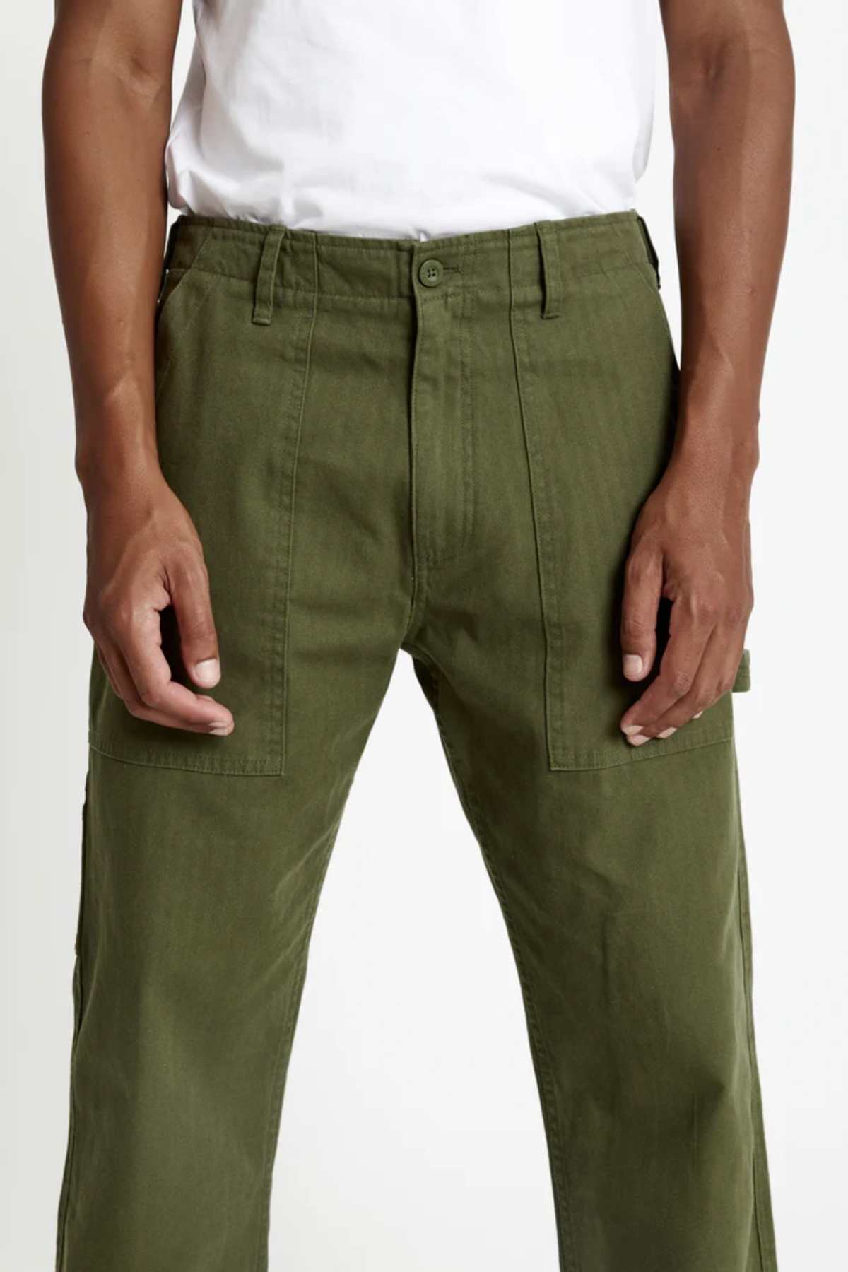 A person wearing the Mr Simple Carpenter Pant in olive green with large front pockets and a white t-shirt, shown from waist to mid-thigh. The workwear-inspired design combines style and functionality.