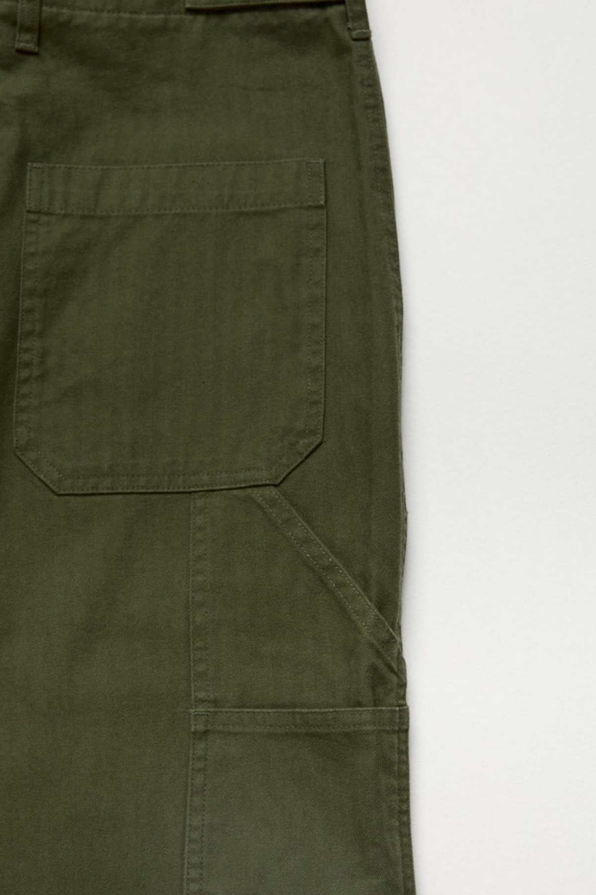 Close-up of the Mr Simple Carpenter Pant in olive green, crafted from 100% cotton with visible stitching, a large rectangular pocket, and a seam detail on the right leg, set against a light background.