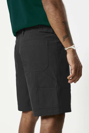 A side view shows a person in Mr Simple Carpenter Walk Short, featuring relaxed fit dark gray cargo style, and a green shirt. A tattoo adorns their arm and they wear a white string bracelet. The plain light background highlights the casual ensemble's herringbone cotton fabric.