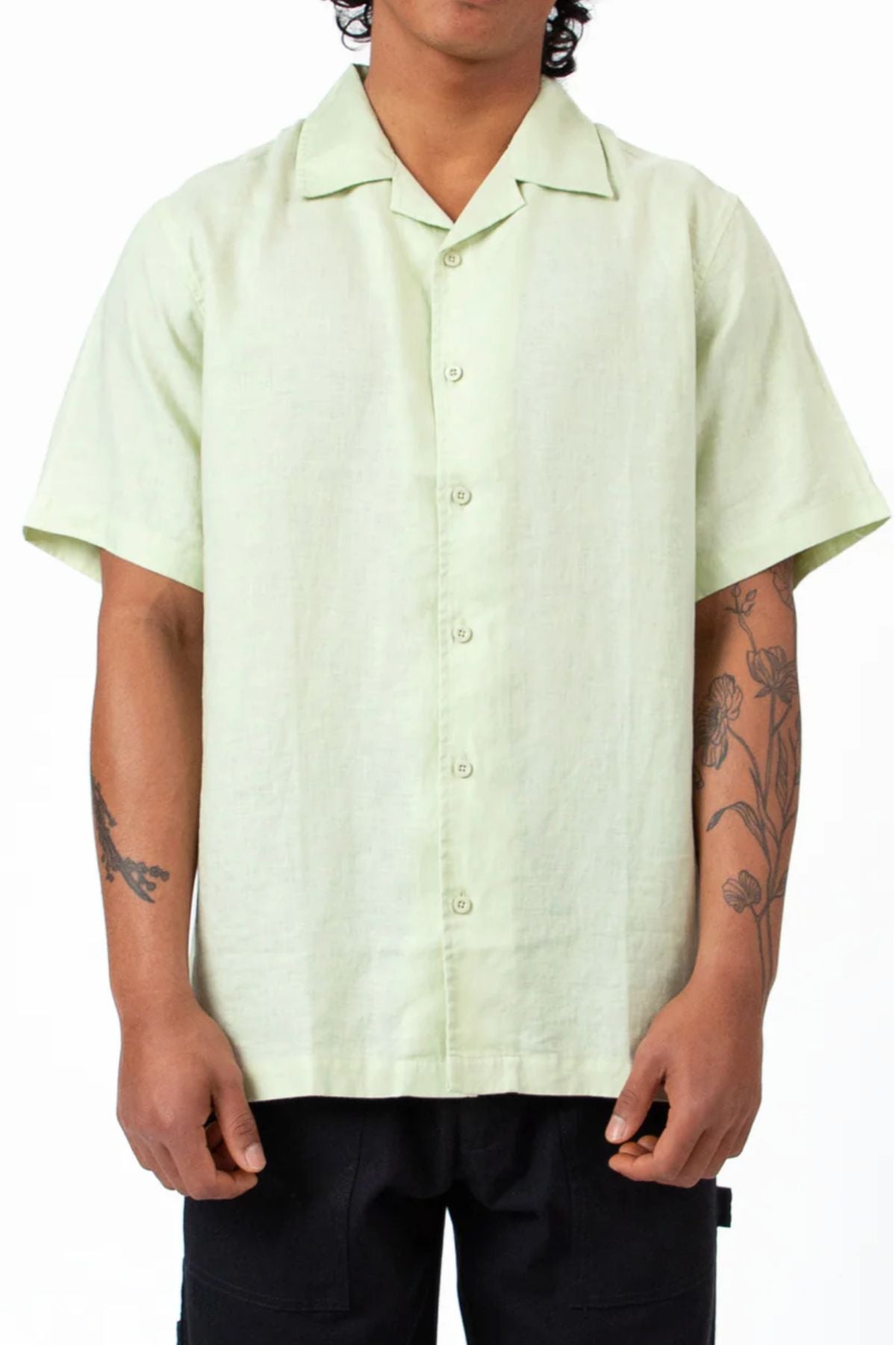 A person wears the Mr Simple Cuban Linen Short Sleeve Shirt in light green with black pants, standing with relaxed arms. Flower and branch tattoos are visible on both forearms against a plain white background.