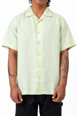 A person wears the Mr Simple Cuban Linen Short Sleeve Shirt in light green with black pants, standing with relaxed arms. Flower and branch tattoos are visible on both forearms against a plain white background.