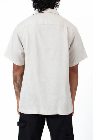 Seen from the back, a person with short, curly hair wears the Mr Simple Cuban Short Sleeve Shirt in a light color and black pants, displaying tattoos on both arms against a plain white background.