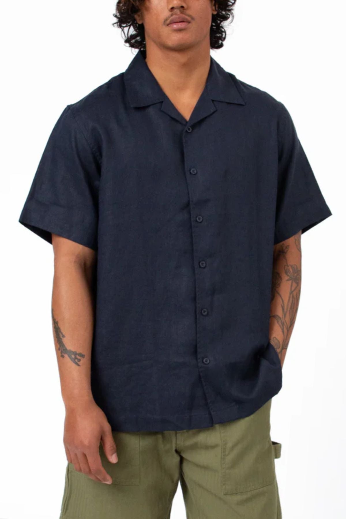 A person with curly hair wears a relaxed fit, dark navy Mr Simple Cuban Short Sleeve Shirt and olive green pants, standing with one hand in their pocket against a plain background. Tattoos are visible on both forearms.