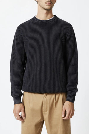 A man wears the Mr Simple Fisher Knit, a relaxed fit black crew neck jumper, with tan pants against a white background. His face is partly visible and his hands rest at his sides.