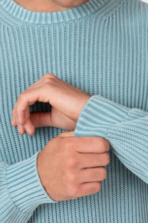 Wearing a light blue Mr Simple Fisher Knit, a person adjusts their sleeve, showing only their lower face and hands. The image highlights the sweater’s relaxed fit and effortless casual style.