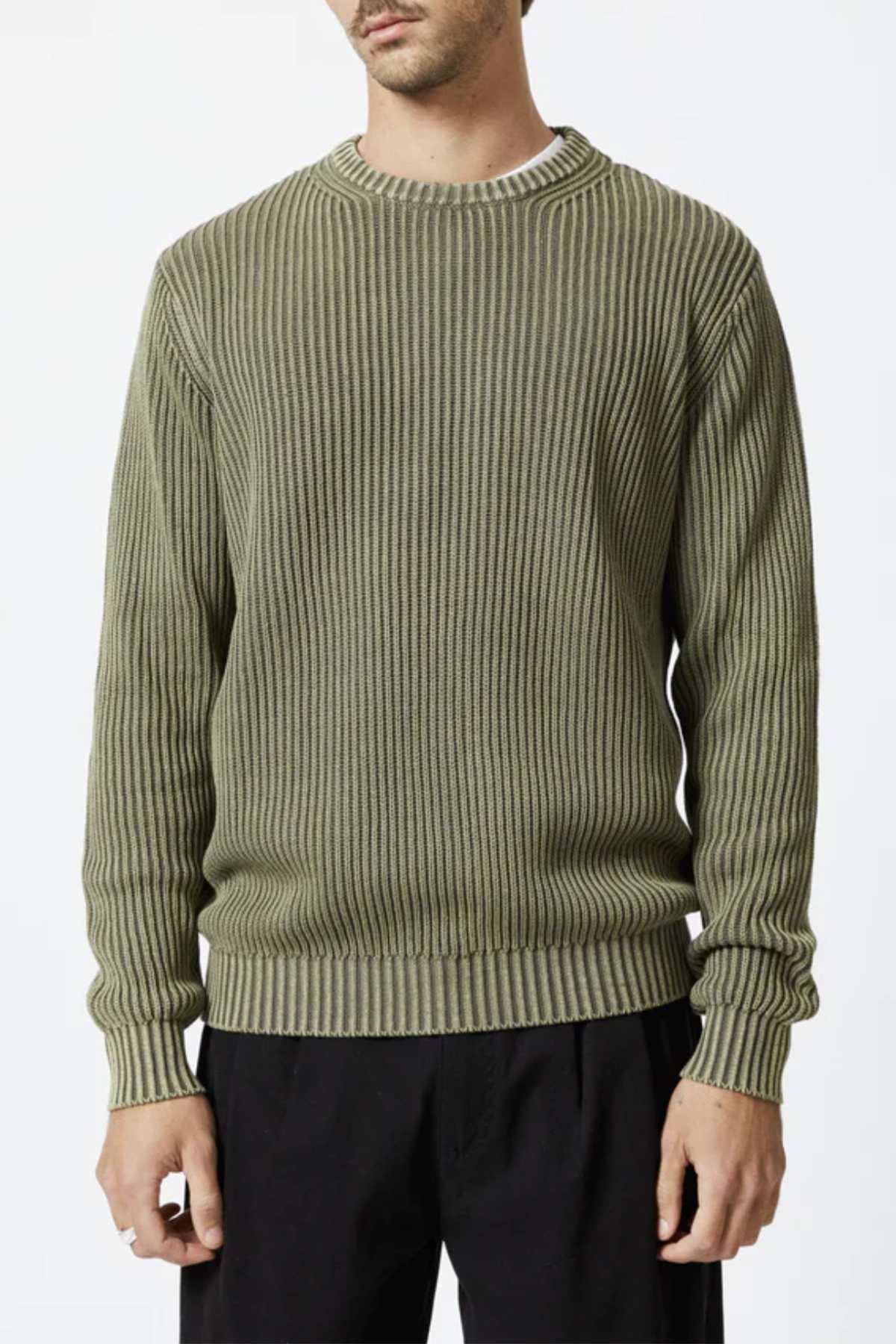 An individual wearing the Mr Simple Fisher Knit sweater in olive green, featuring vertical ribbing, is standing facing forward. The organic cotton sweater boasts a round neckline, long sleeves, and a relaxed fit. The person pairs this with black pants and has a white shirt subtly showing at the neckline.