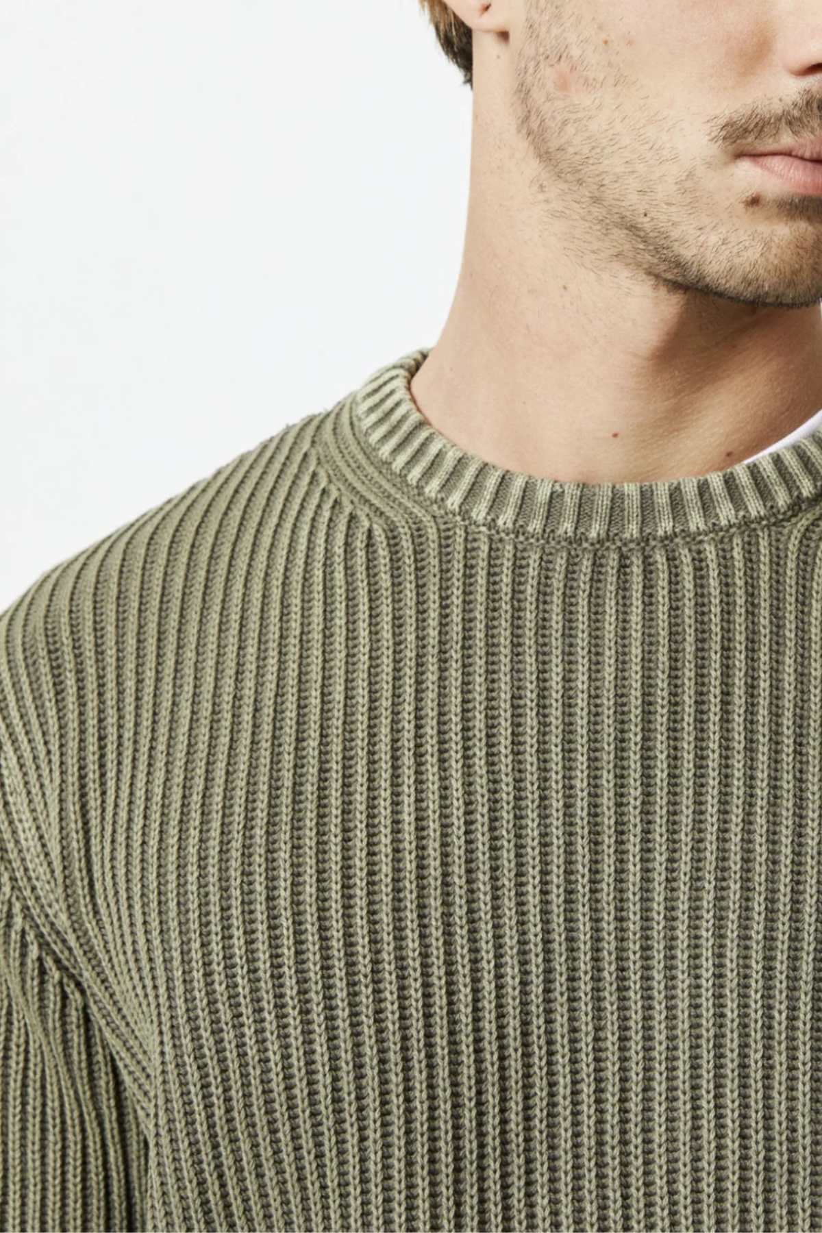 Close-up of a person wearing an olive green Mr Simple Fisher Knit sweater. The focus is on the upper half of the torso, highlighting the texture and design of the organic cotton fabric in its relaxed fit. The background is plain white, with only the lower part of the face and neck partially visible.