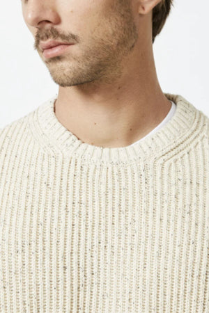 Close-up of a man in a cream Mr Simple Fisher Knit organic cotton sweater over a white shirt, showing his neck, partial face, and mustache against a plain white background.