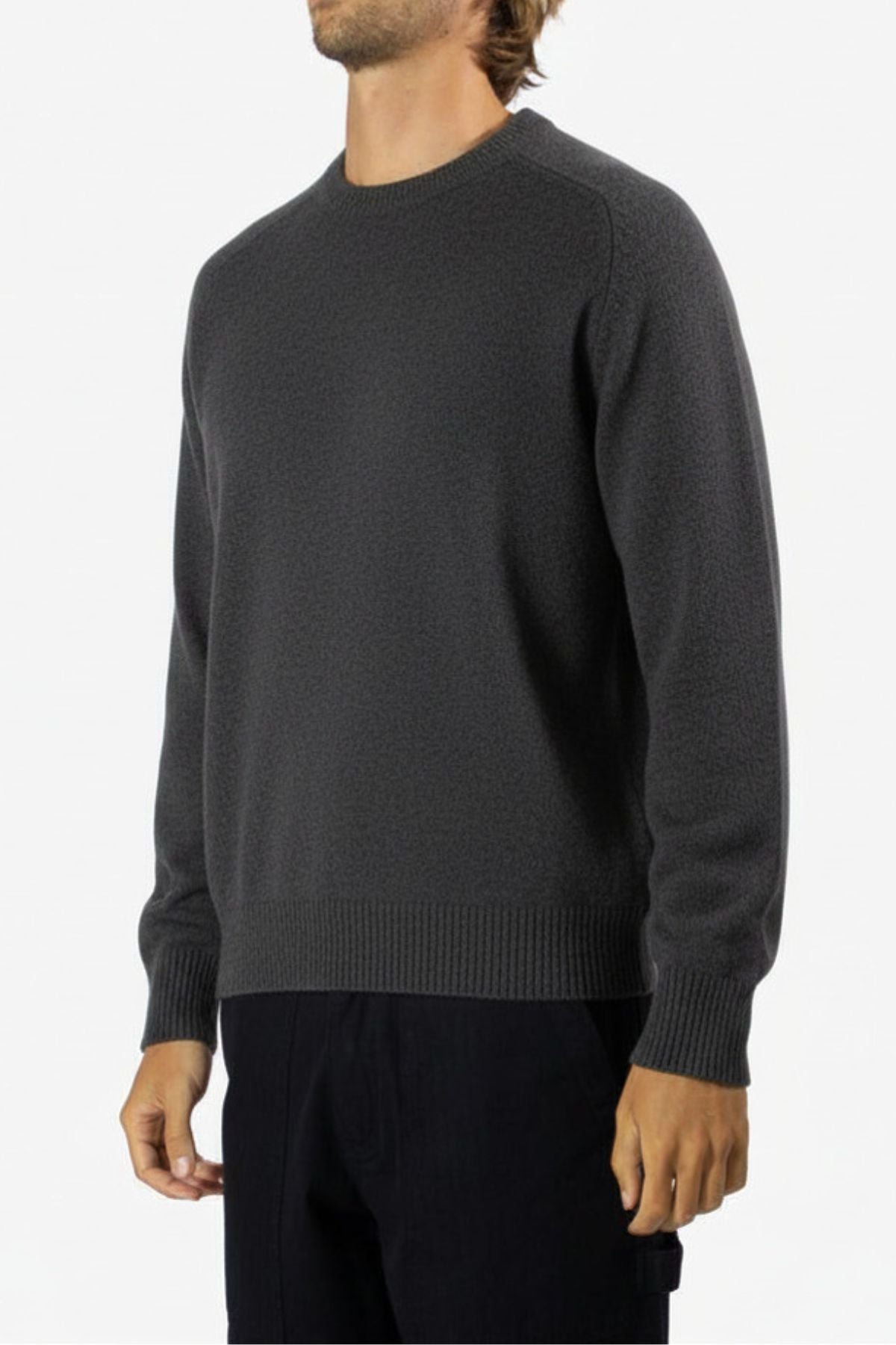 Wearing the Mr Simple Fred Knit in dark gray—a relaxed fit, long-sleeve sweater—paired with black pants, this person stands against a plain light background with their head turned slightly out of frame. Ideal for winter style.
