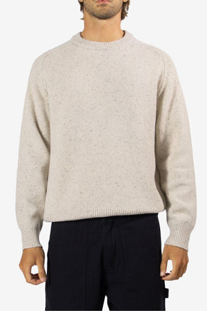 A person wears the Mr Simple Fred Knit—a light beige, long-sleeve 100% wool jumper with ribbed collar, cuffs, and hem—paired with black pants. Their head is partly out of frame against a white background. Ideal for winter wardrobes.