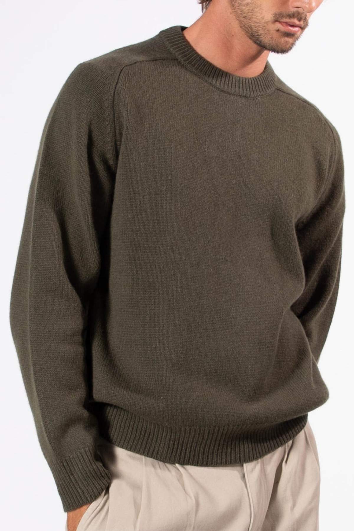 A man is shown from shoulders to mid-thigh, wearing the Mr Simple Fred Knit in relaxed fit olive green, paired with light beige pants, standing against a plain white background.