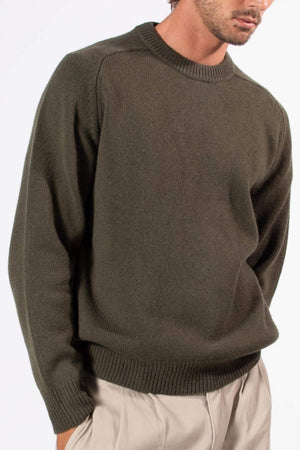 A man is shown from shoulders to mid-thigh, wearing the Mr Simple Fred Knit in relaxed fit olive green, paired with light beige pants, standing against a plain white background.