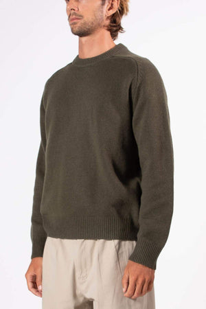 A man wears the Mr Simple Fred Knit, a dark olive green relaxed-fit sweater with a crew neckline and ribbed cuffs and hem, paired with light beige pants, standing against a plain white background—ideal for winter.