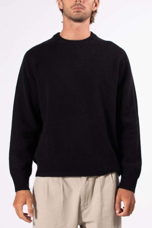 A person wears the Mr Simple Fred Knit, a relaxed fit black wool jumper, paired with light beige pants against a white background—a classic winter wardrobe staple.