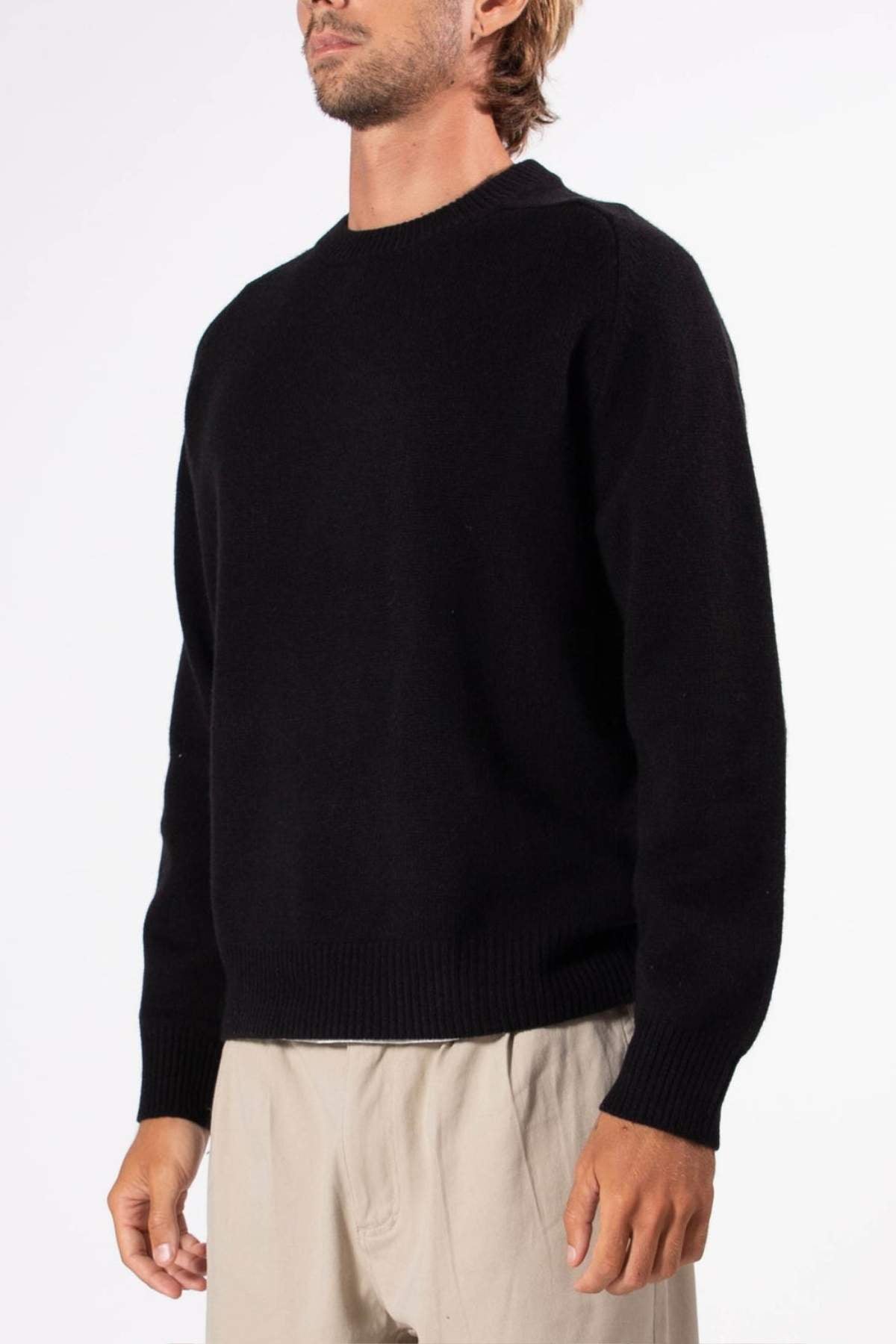 A man wears the Mr Simple Fred Knit black wool jumper with a relaxed fit and light beige pants, standing against a plain white background.