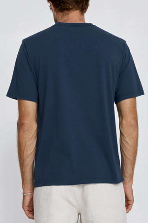 A man is seen from behind wearing the Mr Simple Heavy Weight Tee in a dark blue, boxy loose fit made from 100% organic cotton, paired with light-colored pants. His arms rest at his sides against a plain background.