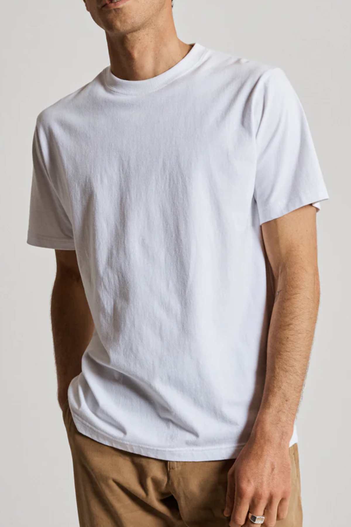 A person models against a neutral backdrop wearing the Mr Simple Heavy Weight Tee, made from 100% organic cotton for ultimate comfort, paired with light brown pants. The image shows their upper body with the face partially obscured and their left hand in their pocket.