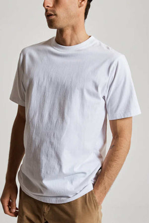 A person in a Mr Simple Heavy Weight Tee (plain white) and light brown pants stands with one hand in their pocket, exuding comfort. Set against a neutral gray background, the focus is on their 100% organic cotton attire, with their face partially obscured.