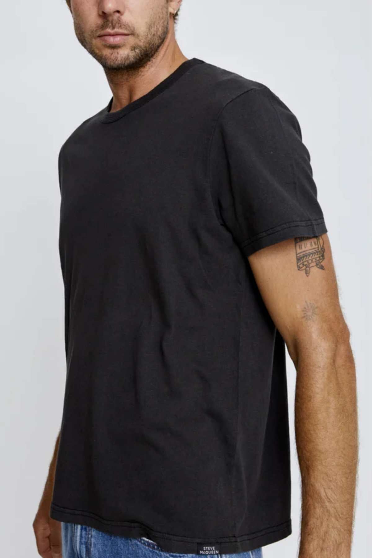 Wearing the Mr Simple Hemp Tee in plain black and blue jeans, a man shows his right forearm tattoo against a neutral light gray backdrop, subtly highlighting his sustainable attire.