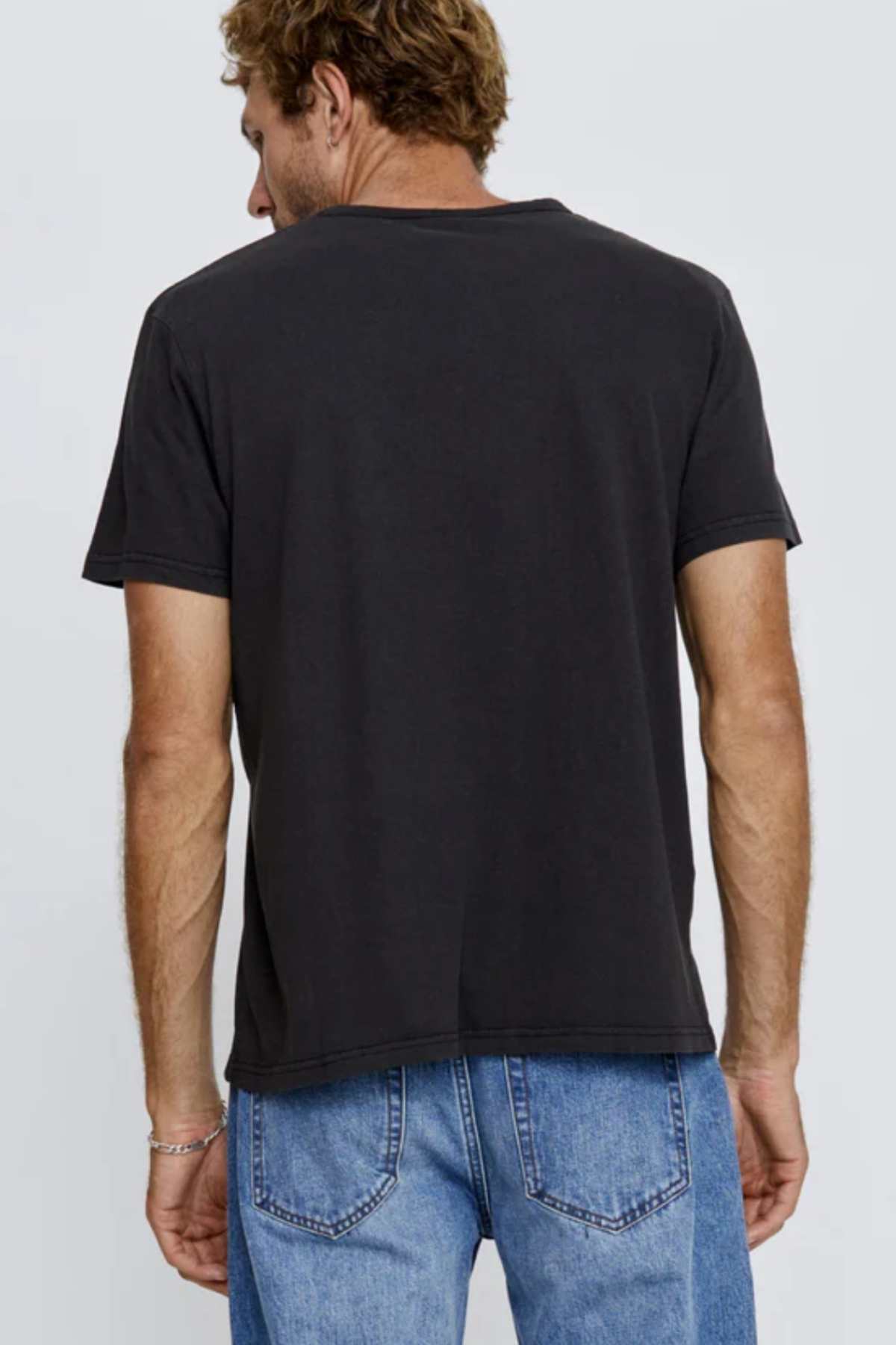 A man with short hair faces away, dressed in a plain black Mr Simple Hemp Tee and blue jeans. His arms rest relaxed at his sides against a minimalist backdrop, embodying a casual yet eco-friendly fashion choice.