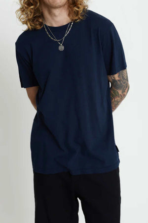 A person with long curly hair is wearing a navy Mr Simple Hemp Tee and black pants, standing with arms behind their back. They showcase a tattoo on their left arm, layered necklaces, and an eco-friendly choice of organic cotton against a plain white backdrop.