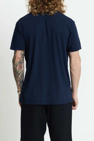 The individual with long curly hair and colorful arm tattoos stands facing away from the camera, wearing a dark blue Mr Simple Hemp Tee made of organic cotton and black pants.