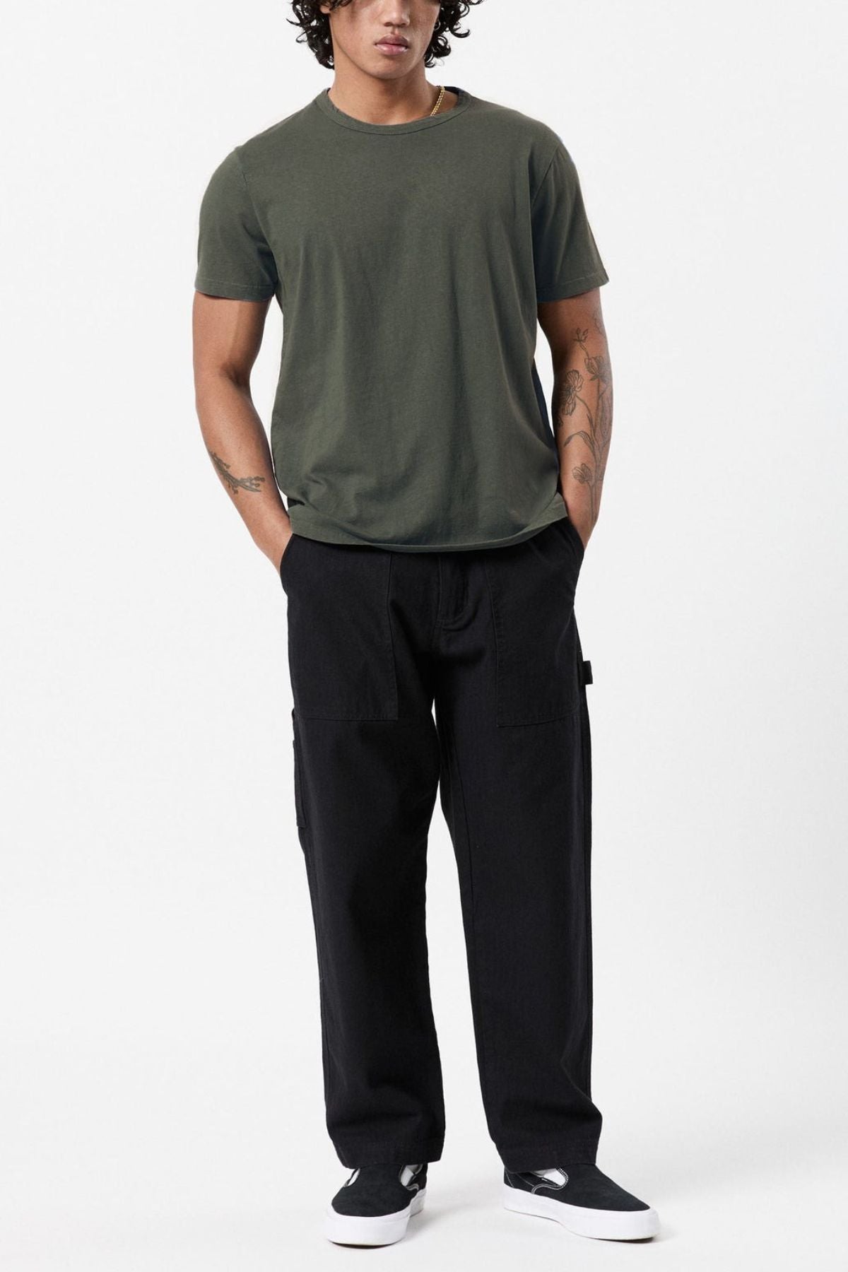 A person stands against a white background wearing the Mr Simple Hemp Tee in dark green and black pants. They have curly hair and visible tattoos on their arms. Their hands are in their pockets, and they are wearing black and white shoes.