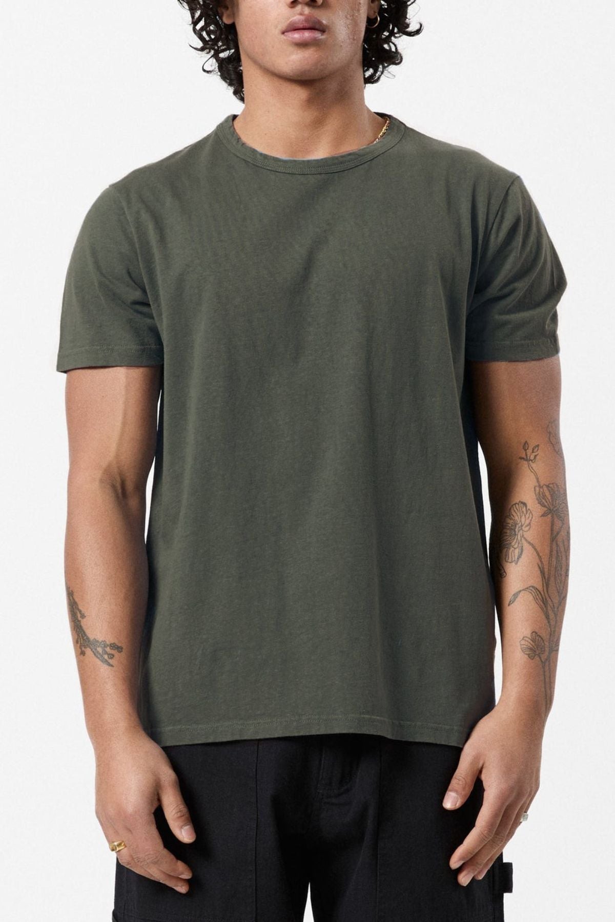 A person in an eco-friendly Mr Simple Hemp Tee in dark green, paired with black pants, stands against a white background. They have curly hair and visible tattoos on both arms, highlighting their commitment to sustainability in fashion.