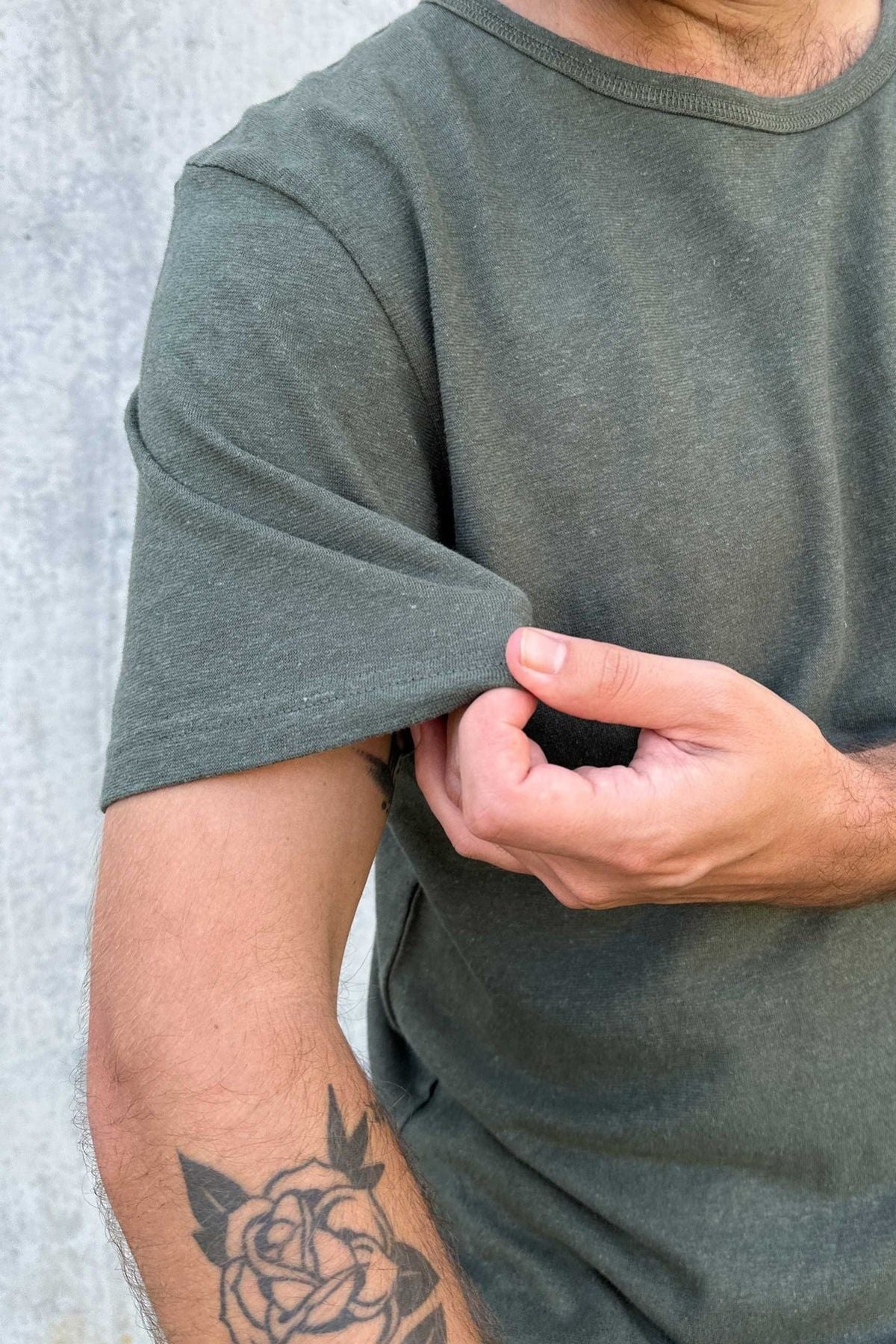 Wearing a Mr Simple Hemp Tee, an individual rolls up their sleeve to reveal various tattoos, including a rose design on their arm. The focus is on the intricate tattoo and the hand adjusting the sleeve against a gray wall backdrop.