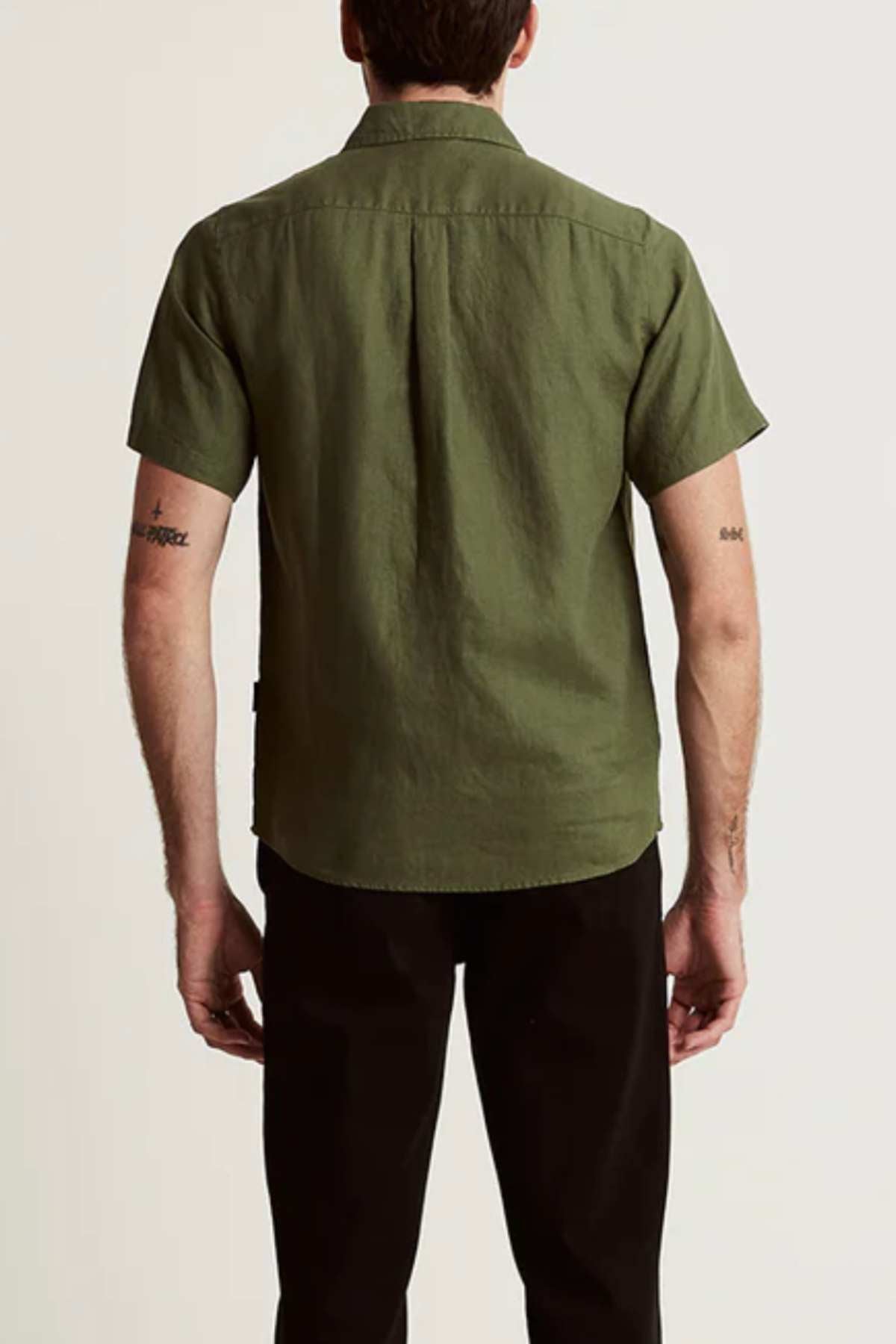 A man stands with his back to the camera, wearing the Mr Simple Linen Short Sleeve Shirt in olive green and black pants. Small arm tattoos are visible. The lightweight shirt is made from linen. The background is plain and light-colored.
