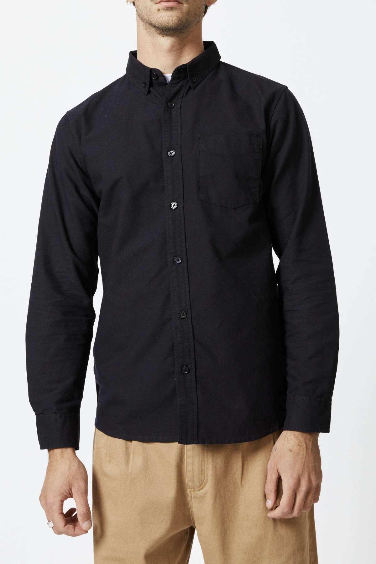 A man wearing a slim fit black Mr Simple Oxford Shirt with a chest pocket and beige pants stands against a plain white background. His head is partially out of the frame.