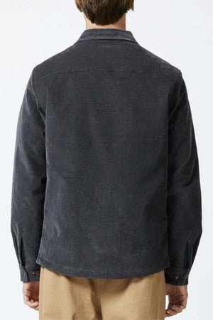 A person with short hair, wearing a Mr Simple Quilted Cord Jacket in dark gray and relaxed-fit beige pants, is standing with their back to the camera. The jacket has buttons on the cuffs and a collar. The background is plain and light-colored.