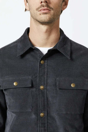 A close-up photo showcasing a person wearing the Mr Simple Quilted Cord Jacket, featuring a dark, relaxed fit with gold buttons and two spacious chest pockets. The cotton corduroy material stands out beautifully against the plain white background. The individual has light facial hair and a mustache, with only the lower part of their face to their torso visible in the shot.