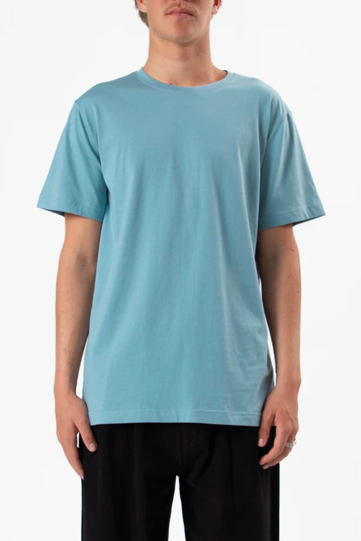 A person models the Mr Simple Reginald Organic Tee in light blue with a comfortable fit and black pants, standing front-facing against a plain white background, cropped at the neck.