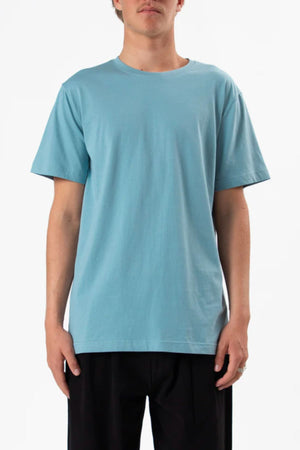 A person models the Mr Simple Reginald Organic Tee in light blue with a comfortable fit and black pants, standing front-facing against a plain white background, cropped at the neck.