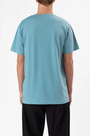 A person stands with their back to the camera, wearing a light blue Mr Simple Reginald Organic Tee and black pants against a plain white background.