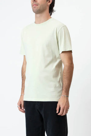 A man wears the Mr Simple Reginald Organic Tee and black pants, standing against a plain white background with his head slightly turned left and arms resting by his sides.